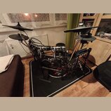 Elektronická bicí souprava umístěná na černém koberci. Sestava obsahuje několik padů, činely a basový pedál s černým rámem. Na spodním bubnu je vidit…