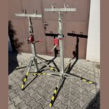 Dvě kovové stojany s nastavitelnou výškou a horními popružími pro uchycení nástroje. Stojany mají stříbrný kovový povrch, červené mechanismy pro nast…
