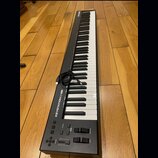 Černý klaviaturní kontrolér s bílými a černými klávesami, položený na dřevěné podlaze. Na levé straně je nápis KEYSTATION 88 MK3 a v horní části logo…