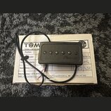 Černý humbucker s šesti viditelnými šrouby v polosovém uspořádání. Na povrchu je rukopisní nápis Tuma. Snímač má připojený černý kabel a leží na bílé…