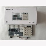 Na fotografii je bílý modul Rhythm Sequencer RS-9 od značky behringer, umístěný před svým původním kartónovým obalem. Modul má bílé provedení s černý…