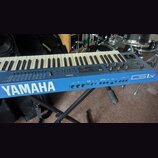 Modrý synthesizér s klaviatura umístěný na černém stojanu. Na přední straně je bílý nápis YAMAHA a vpravo označení modelu CS1x. Na ovládacím panelu j…