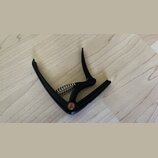 Černý kytarový kapotastán z kovového materiálu s pružinou. V místě spoje je vidět malý červený kruhový prvek. Na předmětu nejsou žádné čitelné značky…