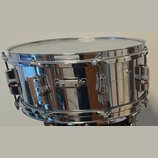 Kovový snare drum s chromovaným, vysoce lesklým povrchem. Na kotečích jsou viditelné loga ve tvaru stylizovaného písmena „S“. Nástroj vykazuje drobné…