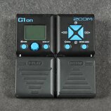 Černý kytarový procesor Zoom G1on s LCD displejem a modrým ovládacím knoflíkem. Na přístroji jsou viditelné nápisy G1on, HOME, ENTER, ZOOM, MENU a RH…