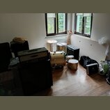 Fotografie zachycuje interiér místnosti s hudebním vybavením. Je zde vidět zlatá bubnová sada, kytarový zesilovač s nápisem "Super", černý basový kom…