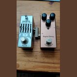 Dvě efektové pedály umístěné vedle sebe na dřevěném povrchu. Levý pedal je stříbrný s nápisy VOLUME, BASS EQ a značkou Amuzik. Pravý pedal je růžozla…