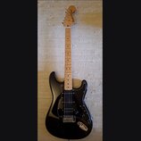 Černá elektrická kytara s jasným dřevěným hmatníkem a bílým pickguardem. Na hlavě je nápis Squier STRATOCASTER. Kytara má tři snímače, pětipositionní…