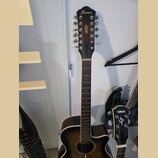 Hnědá akustická kytara s 12 strunami. Na hlavě je zlatý nápis Ibanez a pod ním zlatý symbol šipky. Kytara má tmavý hnědý povrch s výrazným lemováním…