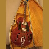 Polodupatá elektrická kytara v červeném provedení s f-otvory, položená v otevřeném kufru s žlutým vnitřkem. Na těle je černý pickguard a kovový ozdob…
