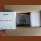 V hnědé kartonové krabici se nachází bílé papírové balení s nápisy HOTONE a ACCESSORY BOX. Vedle něj je tmavý, pravděpodobně plastový předmět zabalen…