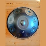 Kulatý kovový handpan s modravým, gradientním povrchem a několika vyhlubecinami (tóny). Nástroj má pletený lem v béžové barvě. V dolní části je bílý…