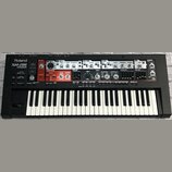 Černý syntetizátor s bílými a černými klávesami. Na levé straně je bílý nápis Roland SH-201 SYNTHESIZER. Horní panel obsahuje množství ovládacích prv…