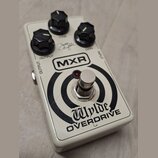 Bílý kytarový efekt MXR Wylde OVERDRIVE s černými oválnými knoflíky. Na horní straně jsou nápisy TONE, GAIN a OUTPUT, vedle nich je viditelný podpis.…