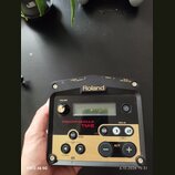 Černý spouštěcí modul s dorém panelu. Na horní straně je bílý nápis Roland a označení konektorů: OUTPUT L/MONO, R, TRIG IN 1, TRIG IN 2 a DC IN PSD A…