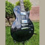 Černá elektrická kytara s tvarem typu Les Paul a hnědým hmatníkem. Na horní části těla je umístěn přepínač s nápisy TREBLE a RHYTHM. Kytara má dva hu…