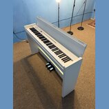 Bílé digitální piano s klasickým provedením a přívodním kabelem. Pod klaviaturou je umístěn černý pedál. Na ovládacím panelu jsou viditelné tlačítka…