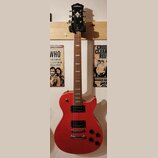 Červená elektrická kytara s tvarem připomínajícím Les Paul, umístěná na nástěnním držáku. Na hlavě je bílý nápis Washburn a logo ve tvaru stylizované…