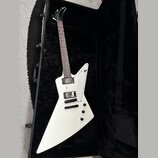 Bílá elektrická kytara s ostrou hranatou formou těla, položená v černém tvrdém kufru. Na hmatníku jsou dva humbuckery a tři ovládací knoflíky, tělo j…
