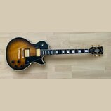 Elektrická kytara s tónem sunburst a černým povrchem na okrajích. Na hlavě je logo Gibson a nápis Les Paul Custom. Nástroj má zlatý hardware, dva hum…
