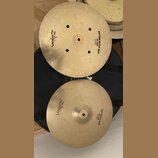 Dva mosazné činely typu hi-hat položené na černém obalu. Horní činel má nápisy Zildjian a BOTTOM QUICK BEAT HI HAT 14"35cm. Dolní činel nese nápisy Z…