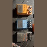 Tři kytarové efekty značky BOSS v kovovém provedení. Horní oranžový pedál má nápis Distortion PS-1, prostřední světle modrý Chorus CEB-3 Bass a spodn…