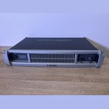 Stříbrný rackový výkonový zesilovač s černým horním krytem. Na předním panelu je nápis PLX 1804 PROFESSIONAL POWER AMPLIFIER a v dolní části logo QSC…