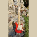 Červená elektrická kytara typu Stratocaster s bílým pickguardem a světlou hnedou hmatníkovou deskou. Na hlavě kytary je viditelné logo Fender. Nástro…