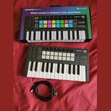 Černý MIDI kontrolér Novation Launchkey Mini s bílými a černými klávesami. Na horní části jsou umístěny šedé pady, ovládací knoflíky a nápis LAUNCHKE…