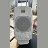 Bílý studiový monitor značky ADAM AUDIO s kovovou mřížkou reproduktoru. V dolní části jsou umístěny ovládací prvky: podsvícené tlačítko s ikonou, vst…