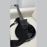 Černá elektrická kytara s tvarem typu Les Paul a bílým lemováním korpusu. Má dva humbuckery, čtyři ovládací knoflíky a černý popruh. Na hmatníku jsou…