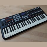 Černý MIDI kontrolér s klaviaturní součástí a bílými a černými klávesami. Na levé straně je nápis AKAI professional MPK49 a mřížka šedých padů. Horní…