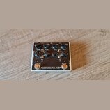 Kytarový efekt pedál v bílém kovovém šasi. Na horní straně je nápis MICROTUBES B7K ULTRA, DARKGLASS ELECTRONICS - MADE IN FINLAND. Pedál má dva spína…