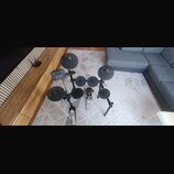 Elektronická bicí souprava s černými padmi a činely na kovovém stojanu. Na ovládacím modulu je vidět nápis YAMAHA a označení DTX. Souprava obsahuje č…