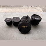 Pět černých textilních přepravních tašek na činely v různých velikostech. Na jedné z tašek je zlatý nápis Zildjian, na jiné bílý nápis ROCKBAG a na d…