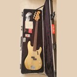 Zlatá elektrická basová kytara v otevřeném černém kufru s červeným nápisem Fender. Na hlavě nástroje je logo Fender. Kytara má hnědý hmatník a dvě sn…