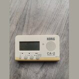 Bílý chromatický ladič značky KORG, model CA-2. Zařízení má digitální displej a mřížku reproduktoru. Na spodní straně jsou tlačítka s nápisy CALIB, A…