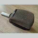 Kovový mikrofon s obdélníkovou mřížkou a stříbrným hrdlem. Na černé liště u mřížky je bílý nápis AKG D12E. Povrch mřížky vykazuje drobné známky opotř…