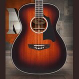 Akustická kytara s tělem v barvě sunburst (přechod od tmavě hnědé k oranžové). Na ozvučovací desce je černý plecháč a v otvoru zvukovníku je vidět ná…