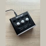 Černý kompaktní přístroj značky behringer s třemi bílými otočnými knoflíky. Na spodní straně jsou viditelné nápisy MIC/LINE GAIN 1, INST GAIN 2 a OUT…