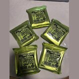 Pět uzavřených balení kytarových strun v zelených fóliových sáčcích. Na každém balení je černý štítek s nápisy ERNIE BALL REGULAR SLINKY, CUSTOM GAUG…