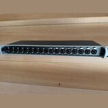 Šedý rackový audio přístroj s černým horním krytem. Na zadním panelu jsou umístěny konektory XLR a dva konektory Ethernet s nápisy SEND a RETURN. V l…