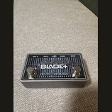 Kytarový pedál v kovovém šedém šasi s černým horním panelem. Na panelu je bílý nápis SWITCH BLADE+ a logo electro-harmonix. Jsou zde vidět dva stoupa…