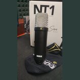 Černý kondenzátorový mikrofon s kovovou síťkou. Na spodní části těla je bílý nápis RØDE. V pozadí je vidět krabice s velkým nápisem NT1 a textem Seri…
