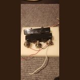 Dva černé elektrické kytarové snímače s nápisem EMG, umístěné na bílém podkladu. K snímačům jsou připojeny tři potenciometry s kovovými hřídelky a ba…
