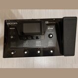 Černý kytarový procesor s karbonovým povrchem a připojeným pedálem značky ZOOM. Na přístroji je nápis G6 Multi-Effects Processor a několik ovládacích…