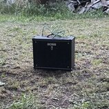 Černý kytarový kombo zesilovač s hranatým tvarem, umístěný na trave. Na přední straně je bílý nápis BOSS a pod ním symbol připomínající stylizované p…