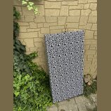 Obdélníkový akustický panel potažený látkou s černobílým geometrickým vzorem. Panel je opřen o žlutou kamennou zeď vedle zelené vegetace. Na povrchu…
