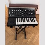 Černý syntetizátor s dřevěnými bočními panely umístěný na černém skládacím stojanu. Na ovládacím panelu je bílý nápis SUBSEQUENT 25 a logo Moog. Nást…