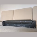 Černý rackový výkonový zesilovač s úchyty po stranách. Na předním panelu je bílý nápis TA600, logo the t.amp a text PROFESSIONAL POWER AMPLIFIER - MK…